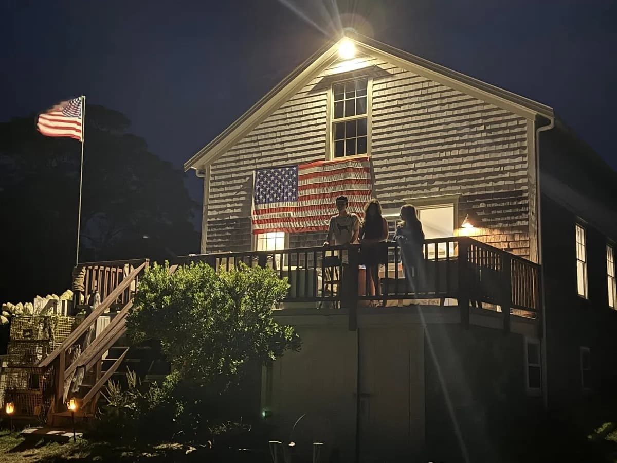 The house at night — American flag on cedar shingles, lobster traps on the porch